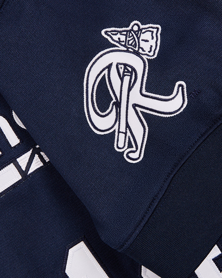 A close-up of navy blue fabric from the Ebbets Field Tom Glavine Richmond Braves V-Neck Jersey shows a white embroidered logo with stylized letters, a flag, and part of a white geometric design and number on this Atlanta Braves garment.