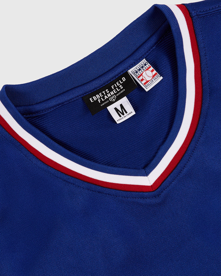 A close-up of the Greg Maddux Peoria Chiefs V-Neck Jersey by Ebbets Field, featuring blue fabric with white and red collar stripes. The inside tag shows 