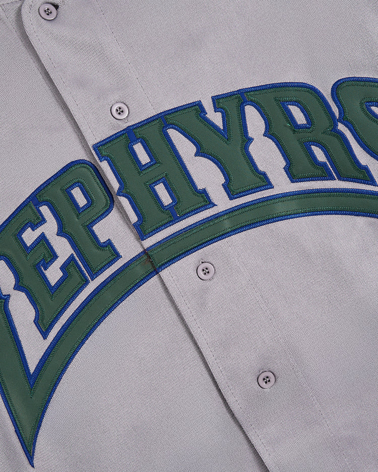 Close-up of the Ebbets Field Barry Larkin Denver Zephyrs Button Up Jersey in grey, showing green and blue embroidered letters spelling part of 