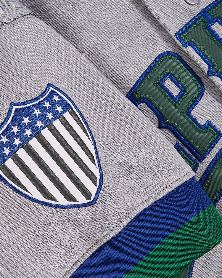 Close-up of a gray Ebbets Field Barry Larkin Denver Zephyrs button-up jersey, showing a stars and stripes shield patch and large green and blue embroidered letters in the background.