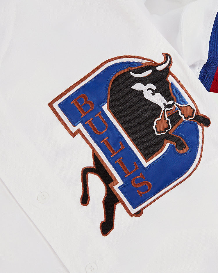 A close-up of the Ebbets Field Chipper Jones Durham Bulls Button Up Jersey shows an embroidered patch with a black bull, blue D, and “BULLS” in red. White buttons are partially visible at the edge of the image.