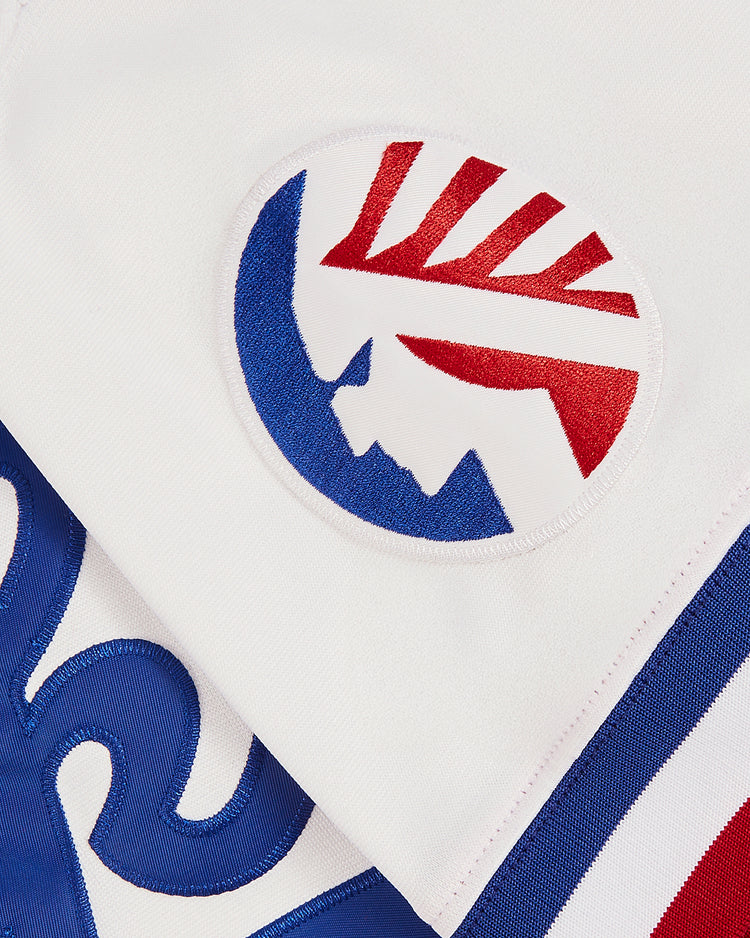 Close-up of the Ebbets Field Tim Raines Memphis Chicks Button Up Jersey shows a white fabric patch with a red and blue mountain logo and sun rays, plus blue swirled and striped designs on the surrounding fabric.