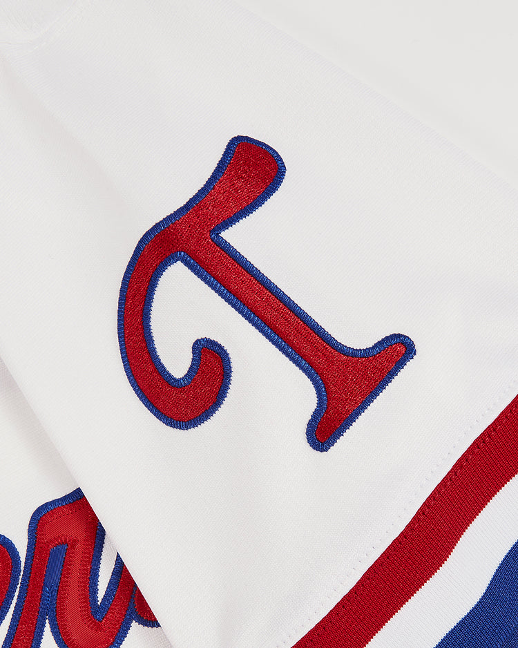 A close-up of the Ebbets Field Pudge Rodriguez Tulsa Drillers Button Up Jersey shows white fabric with a large red and blue outlined letter T stitched on it, plus red, white, and blue stripes at the edge.