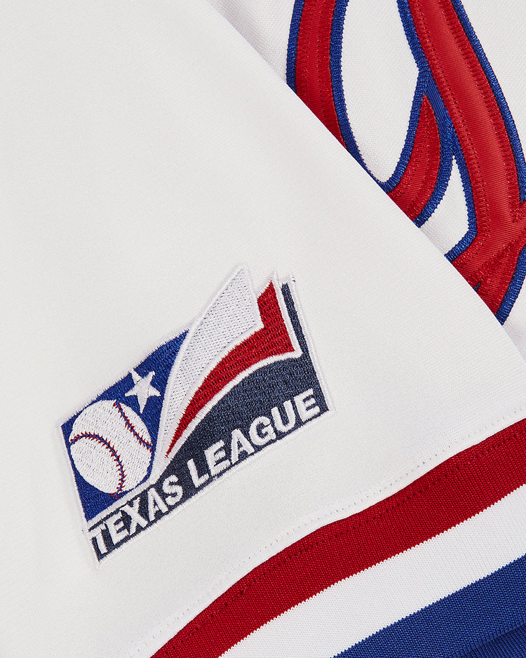 Close-up of a white Ebbets Field Pudge Rodriguez Tulsa Drillers Button Up Jersey with TEXAS LEAGUE patch showing a baseball, star, and red and blue stripes; large Texas Rangers graphic visible in the background.