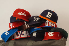 A pile of colorful baseball caps stacked on a bag, each featuring different letters, words, or logos, including Aloha, USA, KC, and various single initials, against a plain background.