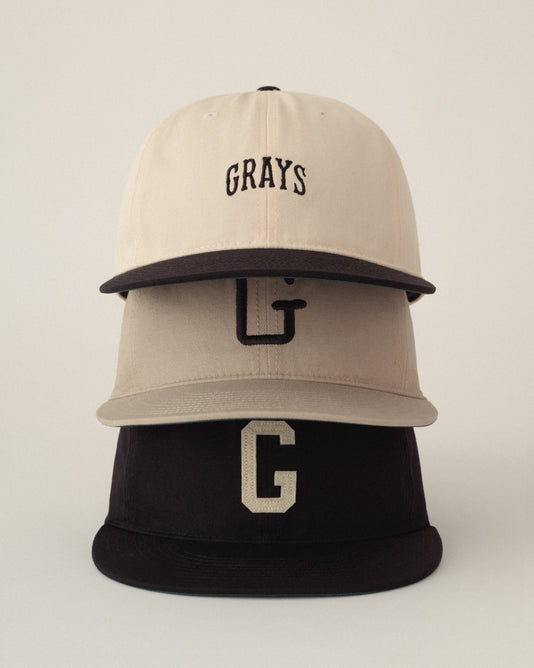 Three baseball caps are stacked vertically. The top cap is beige with GRAYS embroidered on the front, the middle cap is light tan with a large G, and the bottom cap is black with a bold G on the front.