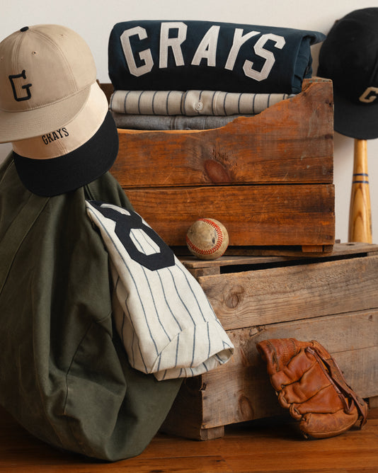 A wooden crate displays vintage-style baseball gear: striped jerseys, a cap with a G, an old baseball glove, a baseball, and a bat, evoking a classic, nostalgic baseball team called the Grays.