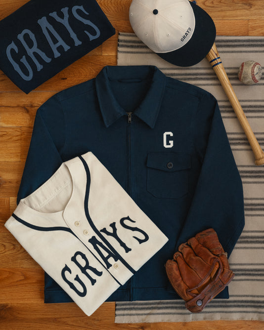 Navy and white baseball apparel with GRAYS lettering, including a jacket, jersey, cap, and folded sweatshirt, are arranged with a glove, bat, and baseball on a striped cloth over a wooden floor.