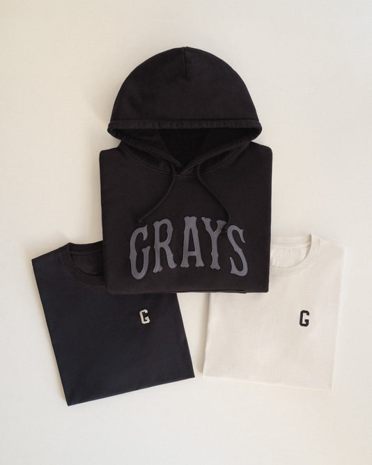 A black hoodie with GRAYS printed on the front is placed above two folded t-shirts, one black and one white, each featuring a small G on the chest.