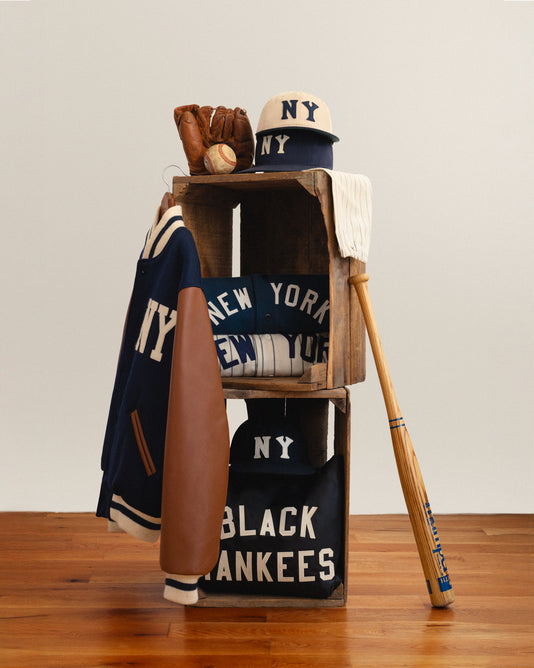 Two stacked wooden crates display NY-themed sportswear, including jackets, shirts, hats, a baseball glove with a ball, and a wooden bat. The items feature NY, NEW YORK, and BLACK YANKEES text.