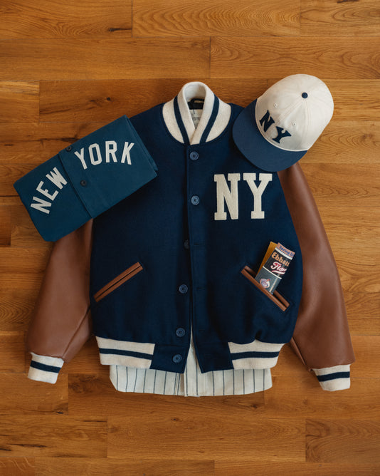 Navy varsity jacket with NY letters, brown leather sleeves, white trim, a folded blue shirt with New York text, a white and navy NY cap, and a pack of chewing gum on a wooden floor.