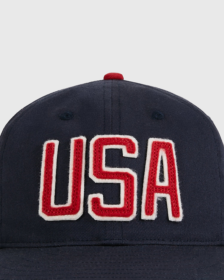 The Ebbets Field USA National Team Cotton Twill Ballcap features bold red “USA” lettering with white outlines on navy blue cotton twill, a red top button, and is displayed on a plain light gray background.