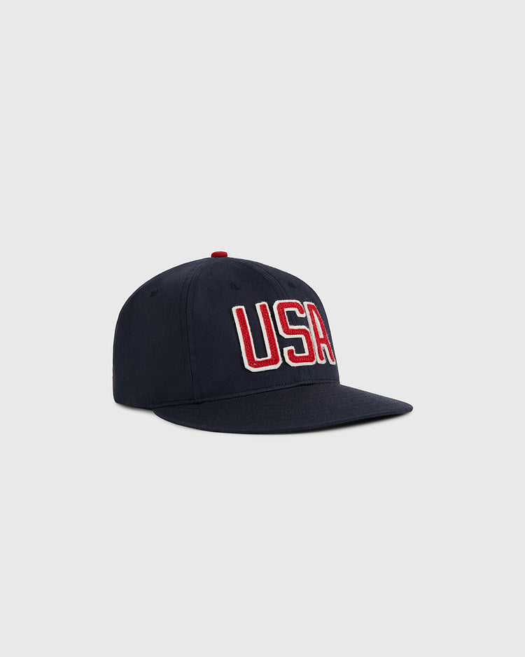 The Ebbets Field USA National Team Cotton Twill Ballcap, inspired by a 1956 Olympic design, features bold red and white embroidery on the front and is shown against a plain light gray background.