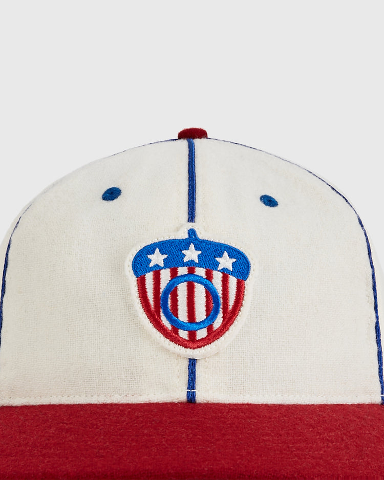 A close-up of the Oakland Oaks 1942 Authentic Wool Ballcap by Ebbets Field, featuring a cream crown, red brim, and a blue and red embroidered acorn logo with stars and stripes.