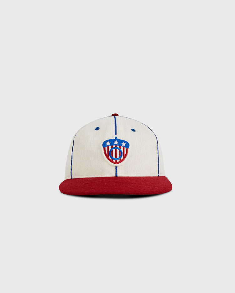 The Ebbets Field Oakland Oaks 1942 Authentic Wool Ballcap features a cream color, blue vertical stitching, red brim, and a front logo with a red, white, and blue shield inspired by the Pacific Coast League’s 1942 acorn and three white stars.