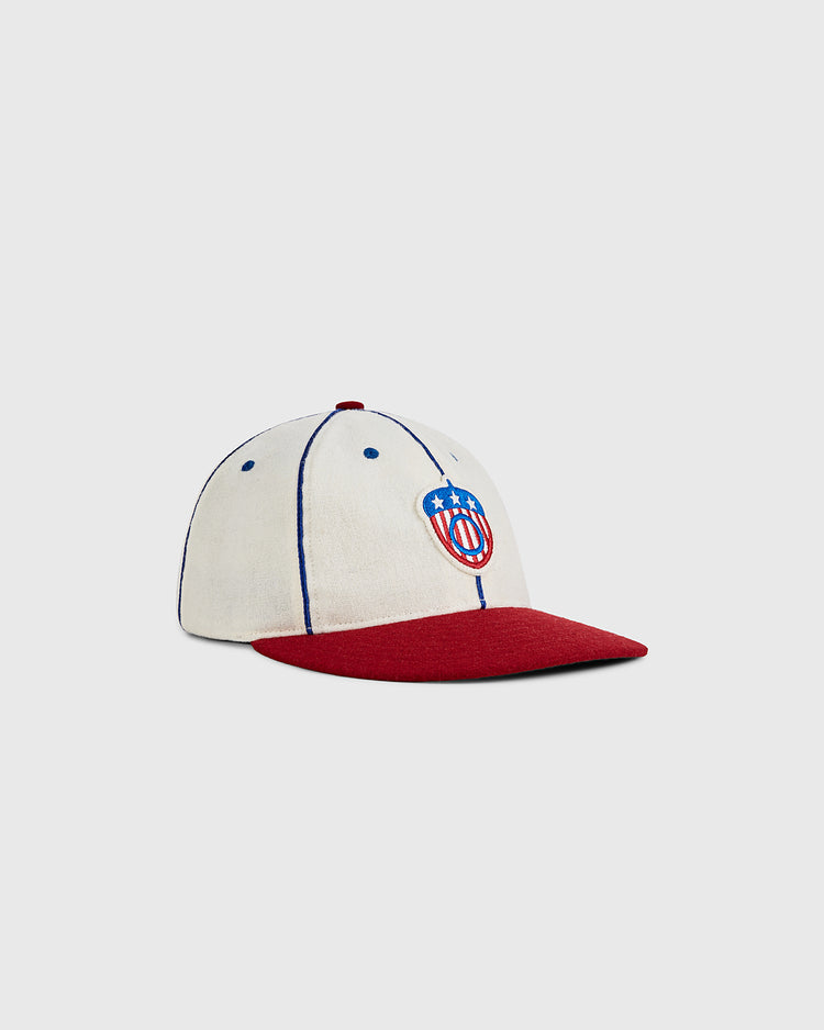 The Ebbets Field Oakland Oaks 1942 Authentic Wool Ballcap features a cream crown, red brim, blue piping, and an embroidered shield logo with blue stars and red stripes, inspired by Pacific Coast League style, shown on a light background.