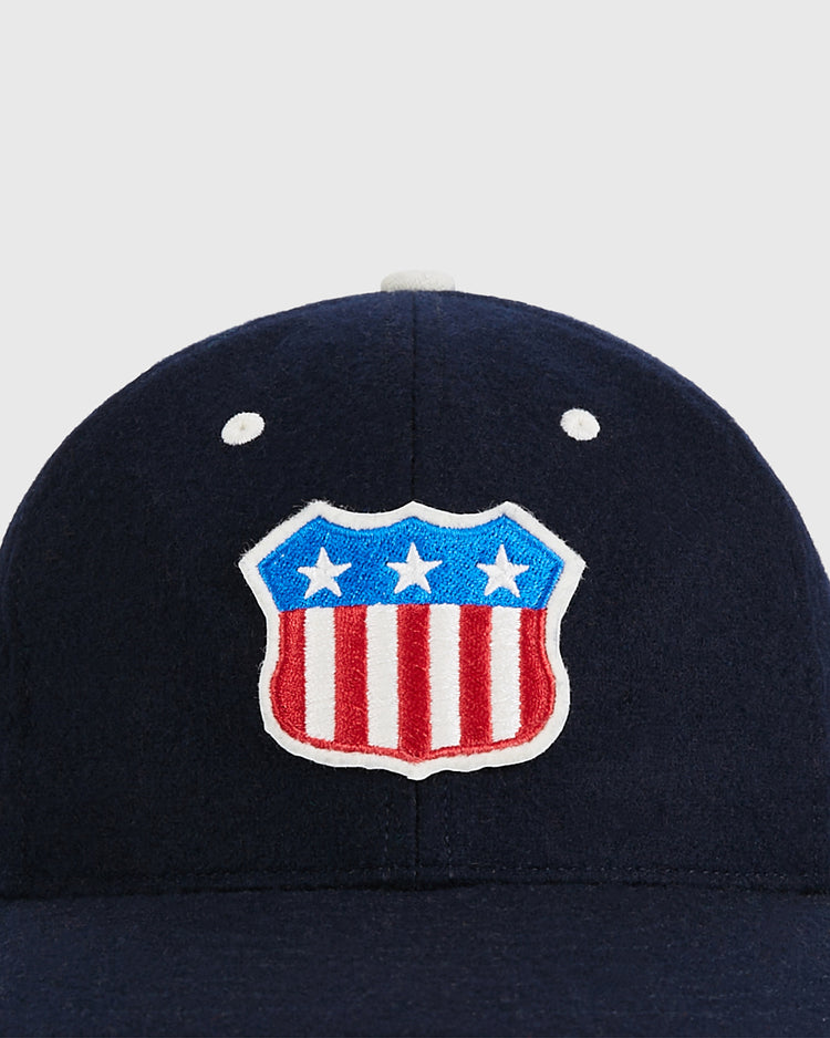 The Ebbets Field USA Stars and Bars Shield Wool Ballcap is a navy blue cap featuring a stars and stripes shield patch on the front, proudly made in the USA.
