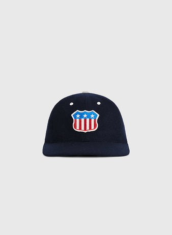The USA Stars and Bars Shield Wool Ballcap by Ebbets Field features a navy blue wool design with a stars and stripes shield patch, displaying three white stars above red and white U.S. flag-inspired stripes on the front. The background is plain light gray.