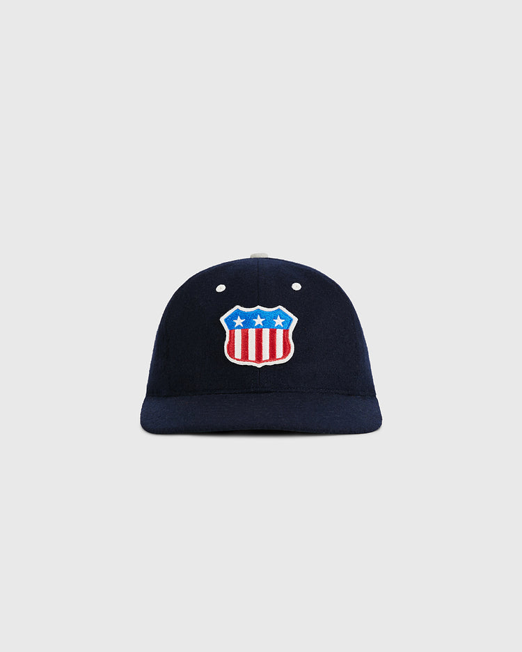 The USA Stars and Bars Shield Wool Ballcap by Ebbets Field features a navy blue wool design with a stars and stripes shield patch, displaying three white stars above red and white U.S. flag-inspired stripes on the front. The background is plain light gray.