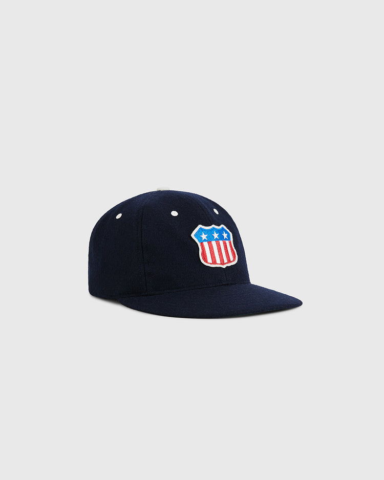 The Ebbets Field USA Stars and Bars Shield Wool Ballcap, in navy blue wool, features a red, white, and blue shield emblem with three white stars on the front, shown against a plain light gray background.