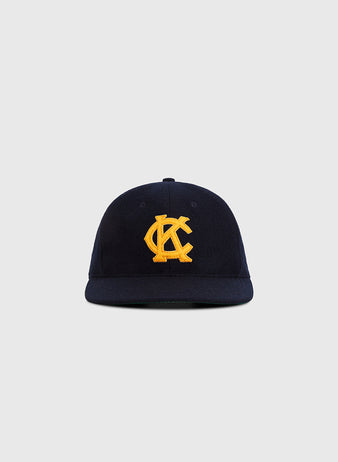The Ebbets Field Kansas City Monarchs 1945 Authentic Wool Ballcap is navy blue with a curved brim and a yellow embroidered overlapping KC logo on the front, shown against a plain light gray background.