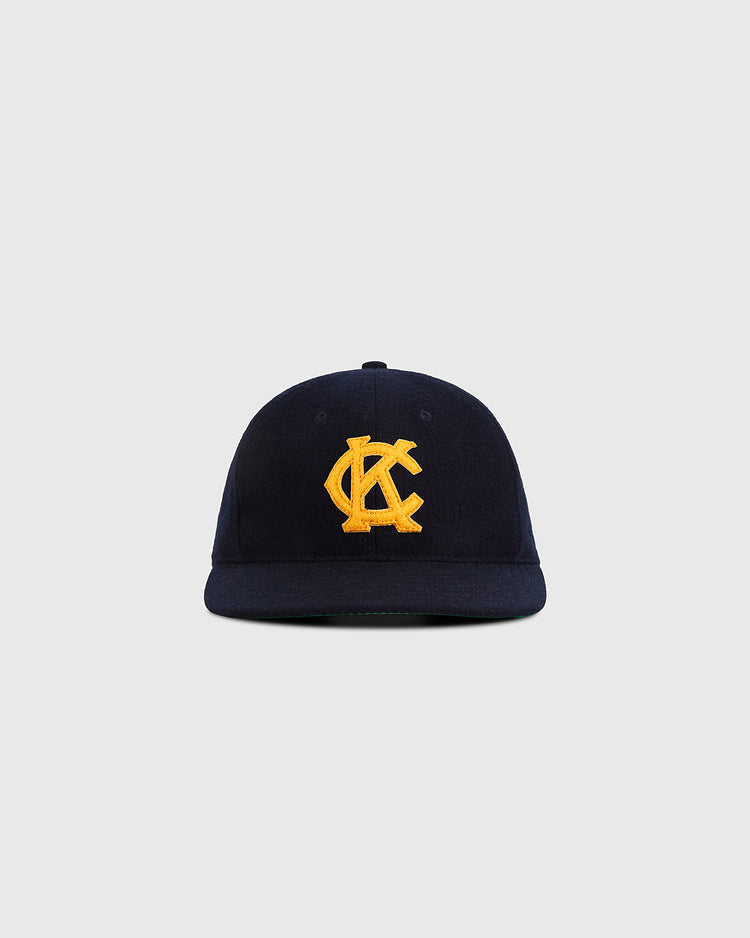 The Ebbets Field Kansas City Monarchs 1945 Authentic Wool Ballcap is navy blue with a curved brim and a yellow embroidered overlapping KC logo on the front, shown against a plain light gray background.