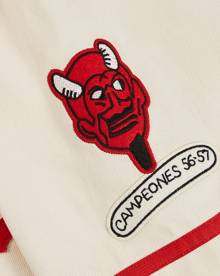 The Ebbets Field Diablos Rojos del Mexico Cotton Twill Baseball Jersey in cream features a red and black devils head logo above a white CAMPEONES 56-57 patch, with red trim accenting the edges.