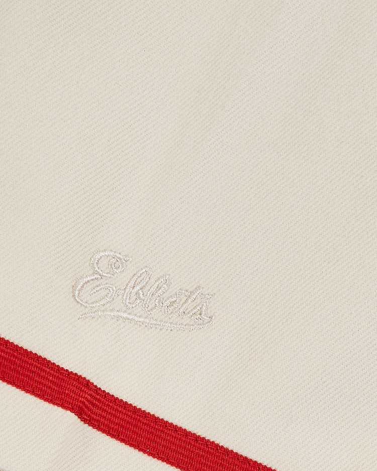 A close-up of cream cotton twill fabric with “Ebbets Field” embroidered in matching thread and a diagonal red stripe, inspired by the 1933 Satchel Paige All-Stars jersey.