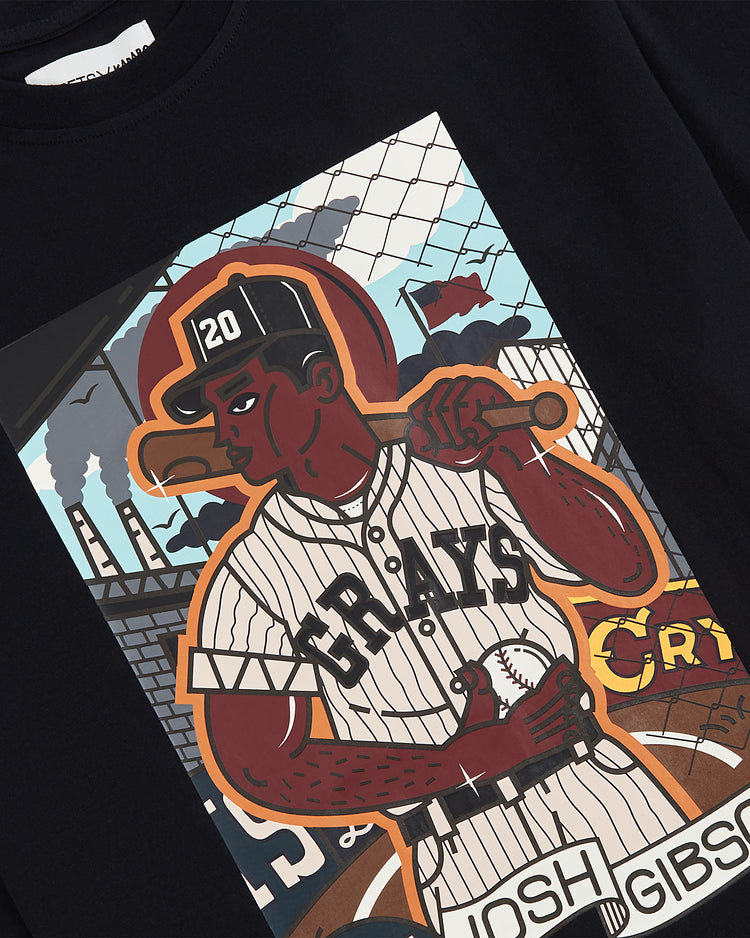 The Ebbets Field Karabo Poppy x Josh Gibson Tee features an illustrated player in a “GRAYS” jersey with a bat and “JOSH GIBSON” ball, set against factories and flags—a tribute to the legend by artist Karabo Poppy.
