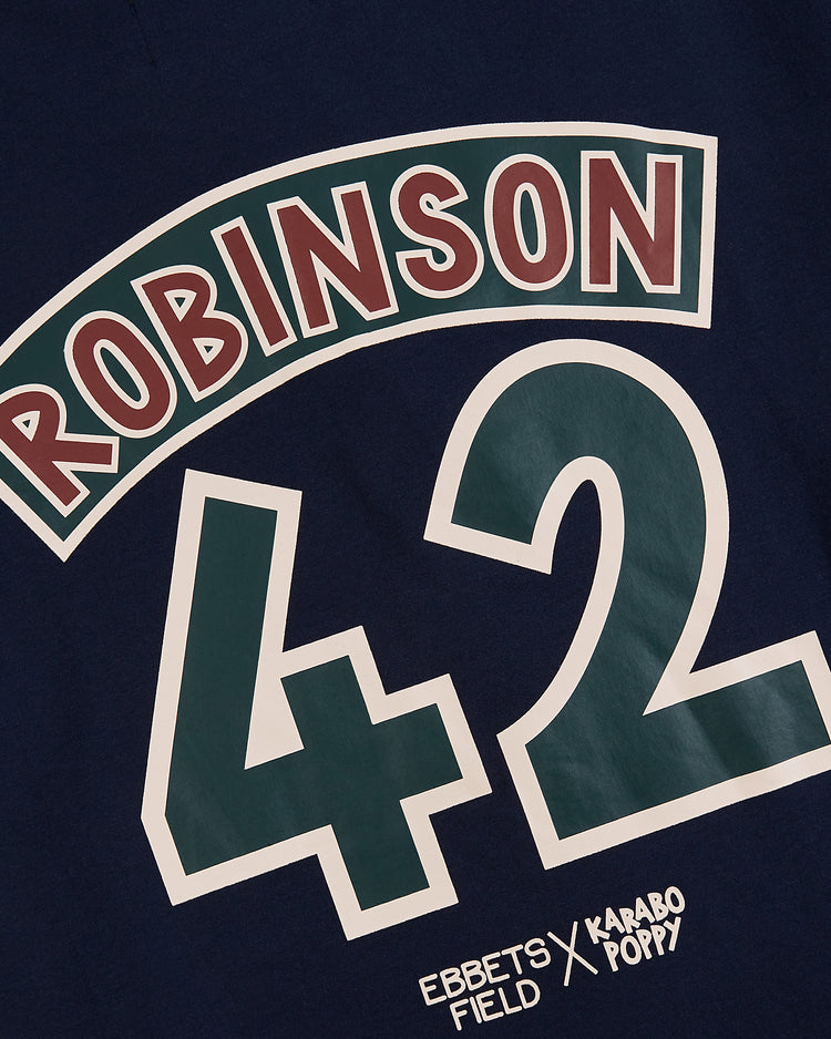 The Ebbets Field Karabo Poppy x Jackie Robinson Tee features navy blue fabric, “ROBINSON” in red and white arching letters, a large green and white 42, and celebrates Black History Month with EBBETS FIELD & KAARO POP! below.