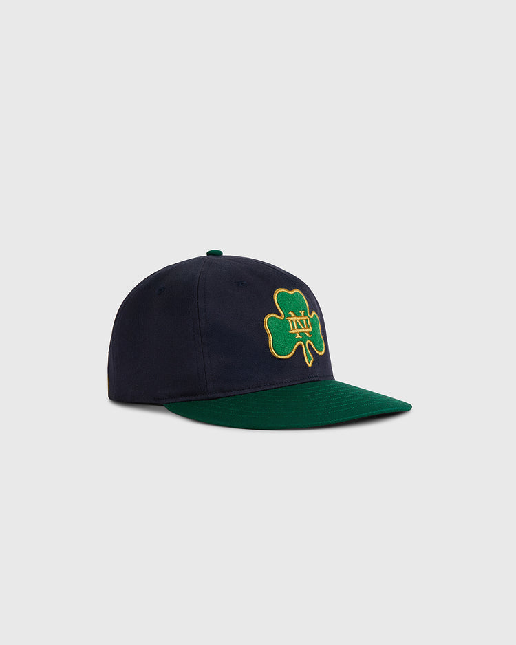 The Ebbets Field University of Notre Dame Shamrock Cotton Twill Ballcap features navy cotton twill, a green brim, a green shamrock outlined in yellow, and a yellow ND logo embroidered on the front, shown on a light gray background.