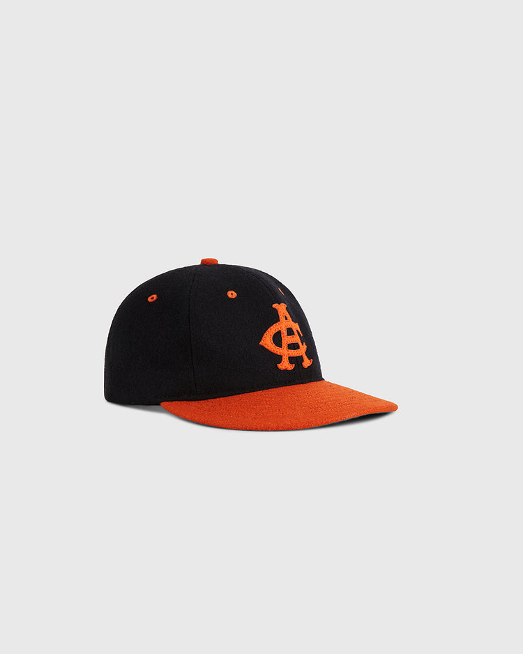 The Ebbets Field Aguilas Cibaenes 1955 Authentic Wool Ballcap features a black crown, orange brim, and an orange “A” logo on the front, all displayed against a plain light gray background.