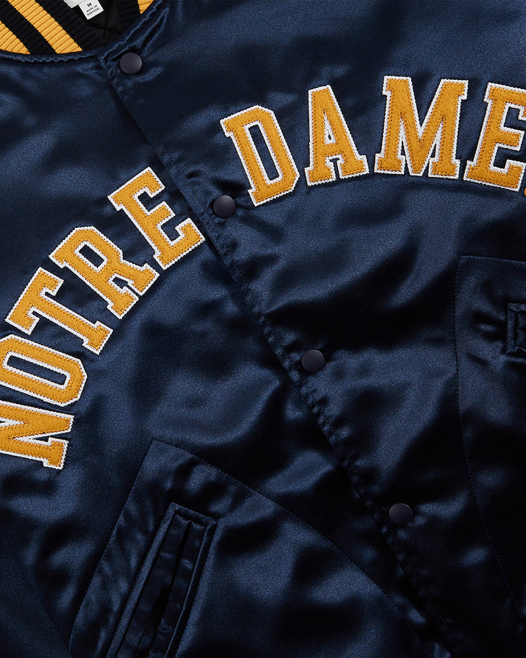 Close-up of the Ebbets Field Notre Dame Satin Varsity Jacket in navy, featuring yellow and white NOTRE DAME lettering, snap buttons, front pocket, and a bold yellow and navy striped collar.