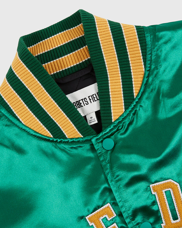 A close-up of the Ebbets Field Notre Dame Satin Varsity Jacket shows a ribbed collar with green, yellow, and white stripes, green snap buttons, and partially visible yellow embroidered lettering on the front.