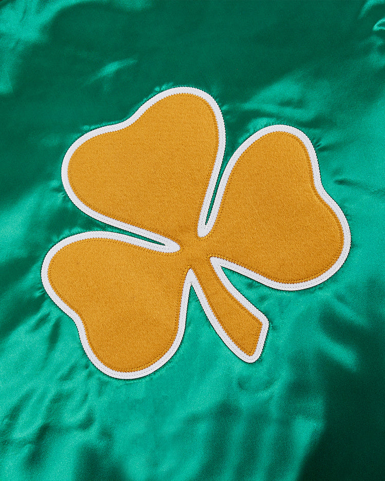 A yellow three-leaf clover with a white outline is sewn onto shiny green fabric, evoking the classic style of the Ebbets Field Notre Dame Satin Varsity Jacket.