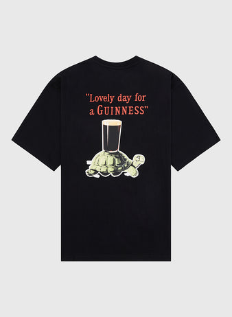 The Ebbets Field Guinness Tortoise Short Sleeve Graphic Tee features a turtle carrying a pint of Guinness below the slogan “Lovely day for a GUINNESS” in bold red and white letters.