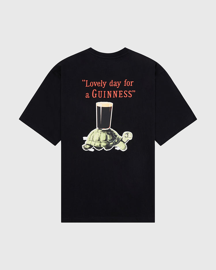 The Ebbets Field Guinness Tortoise Short Sleeve Graphic Tee features a turtle carrying a pint of Guinness below the slogan “Lovely day for a GUINNESS” in bold red and white letters.