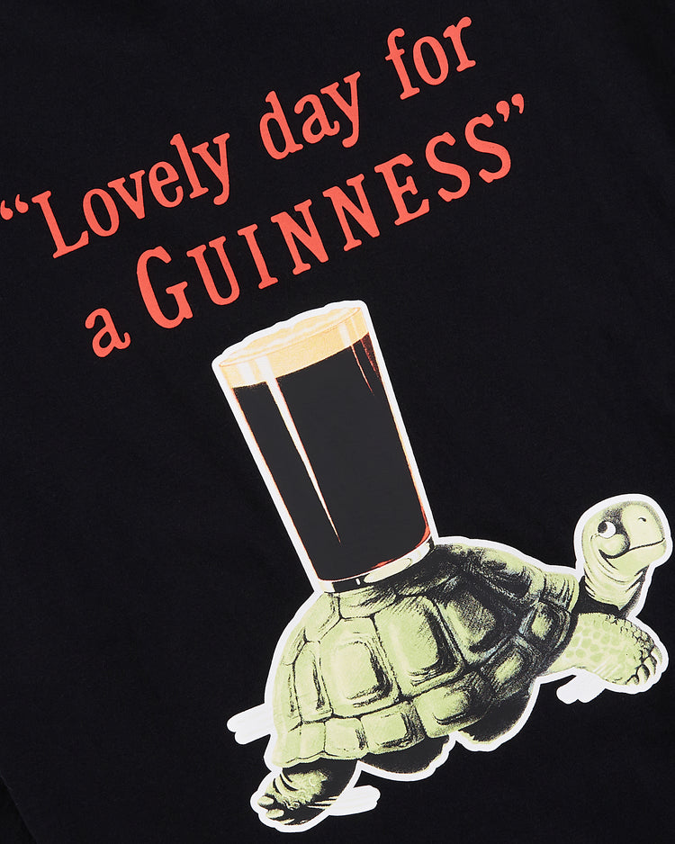 The Ebbets Field Guinness Tortoise Short Sleeve Graphic Tee features a cartoon turtle carrying a pint of Guinness and the phrase “Lovely day for a Guinness” in bold red letters—perfect for fans of unique graphic tees.
