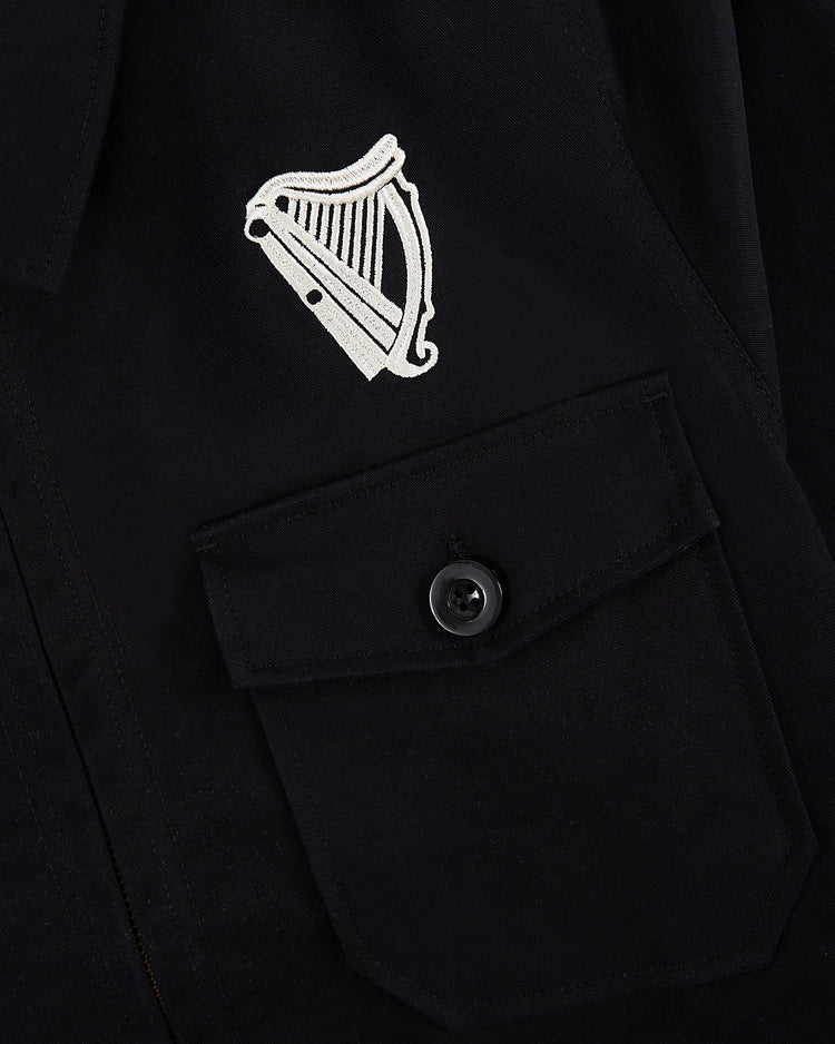 Close-up of the Guinness Cotton Grounds Crew Jacket by Ebbets Field, featuring a white embroidered harp above a buttoned flap pocket on black fabric.