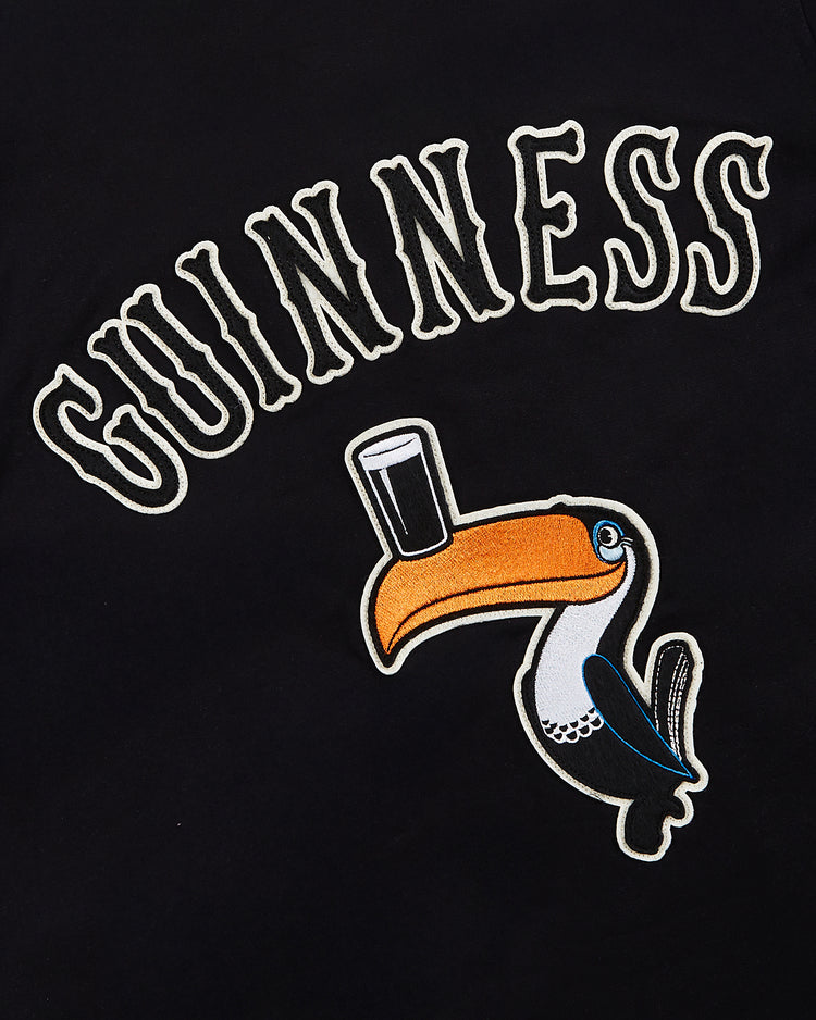 The image shows the word GUINNESS above a toucan balancing a pint of beer on its beak, set against black—a design featured on the Guinness Cotton Grounds Crew Jacket by Ebbets Field.