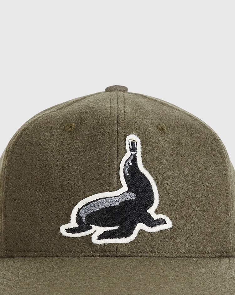 A close-up of the Ebbets Field Guinness Seal Authentic Wool Ballcap in olive green, featuring a black and white embroidered patch of a seal balancing a bottle on its nose.