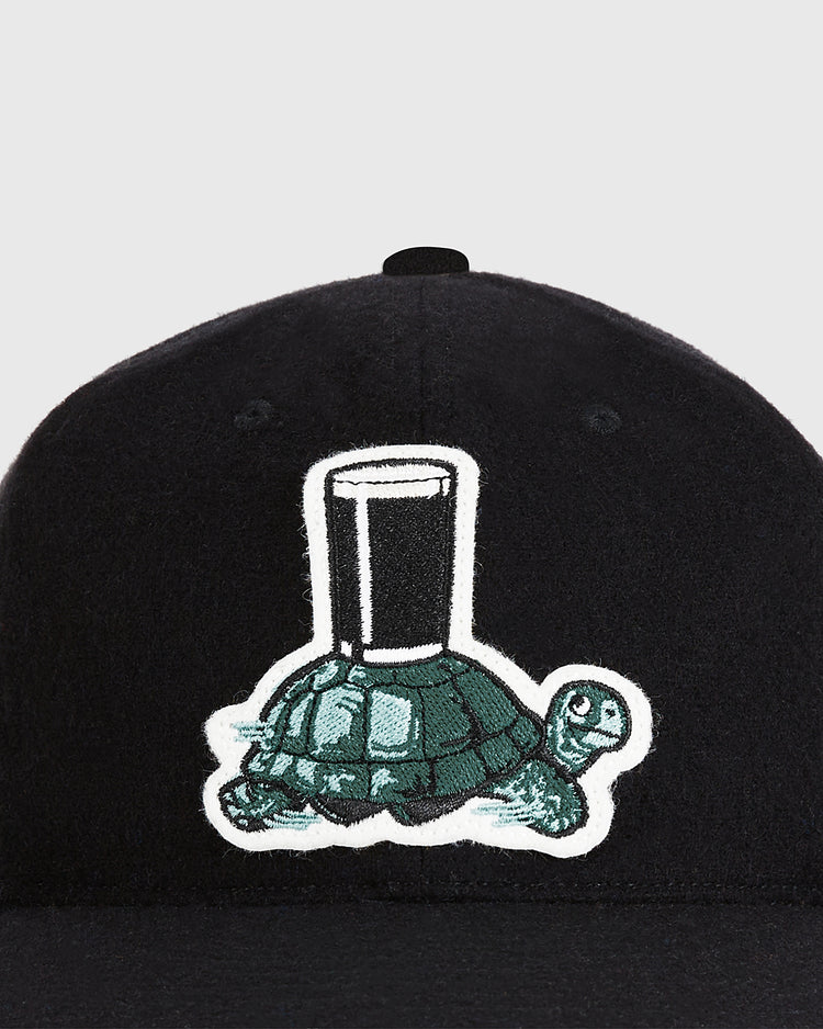 The Ebbets Field Guinness Tortoise Authentic Wool Ballcap is a black hat featuring an embroidered green tortoise carrying a glass of water on its shell.