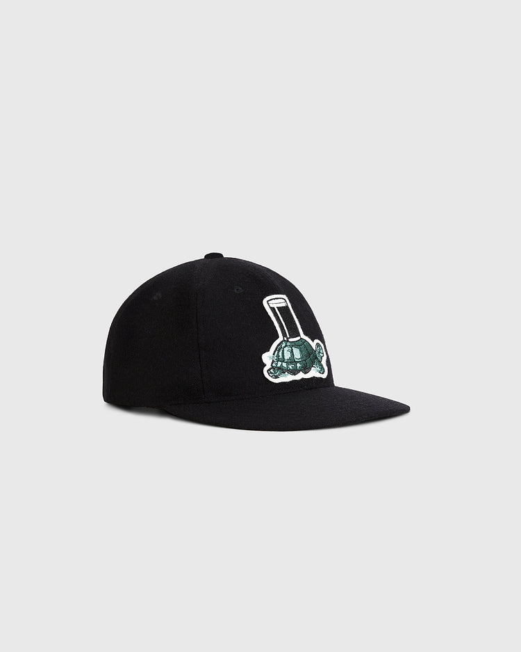 A black Guinness Tortoise Authentic Wool Ballcap by Ebbets Field, inspired by classic styles, features an embroidered green and white lawn mower patch on the front and is shown on a plain light gray background.
