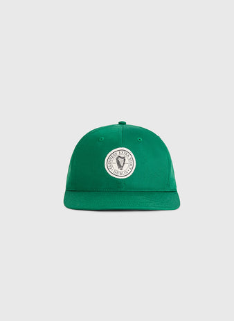 The Ebbets Field Guinness Extra Stout Badge Cotton Twill Ballcap is a green cap with a round white patch featuring a black shield and surrounding text, shown on a plain light gray background.