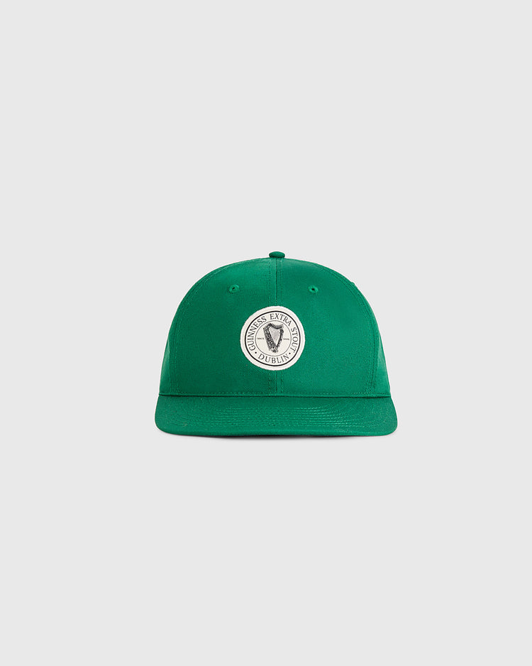 The Ebbets Field Guinness Extra Stout Badge Cotton Twill Ballcap is a green cap with a round white patch featuring a black shield and surrounding text, shown on a plain light gray background.