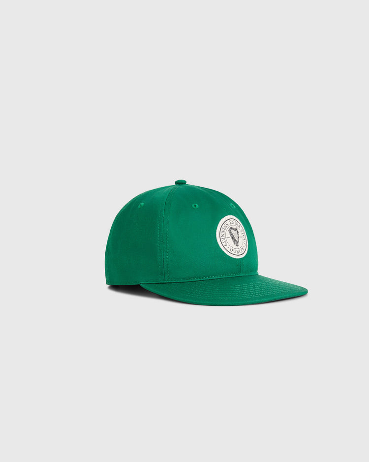 The Ebbets Field Guinness Extra Stout Badge Cotton Twill Ballcap, in green with a round white front patch featuring a graphic and text, is shown at an angle on a light gray background.