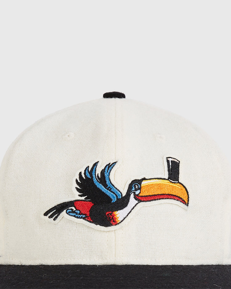 A close-up of the Ebbets Field Guinness Flying Toucan Authentic Wool Ballcap, a white cap with a black brim and a colorful embroidered toucan with outstretched wings and a yellow-orange beak on the front.