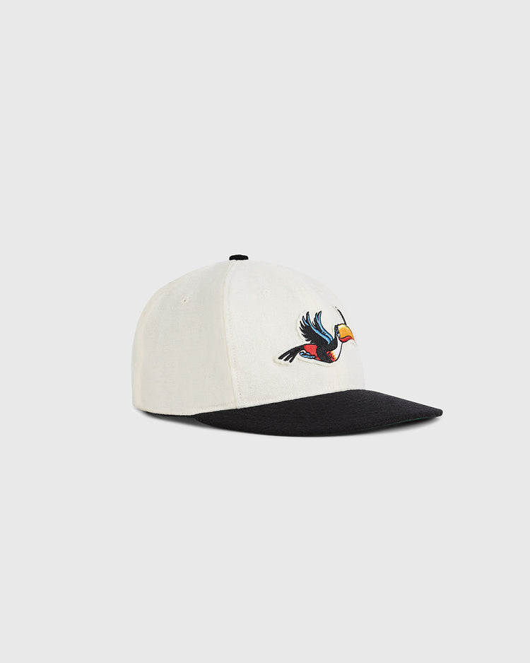 The Guinness Flying Toucan Authentic Wool Ballcap by Ebbets Field is a white cap with a black brim and an embroidered colorful toucan in flight on the front, shown against a plain light gray background.