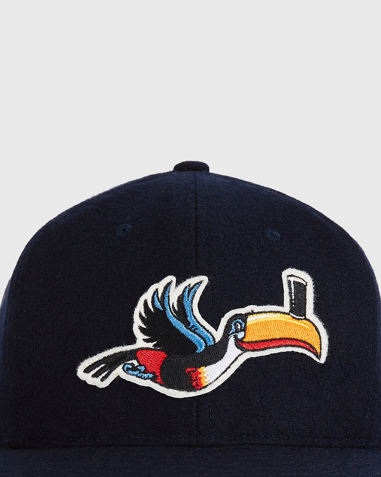 A close-up of the Ebbets Field Guinness Flying Toucan Authentic Wool Ballcap in dark blue, featuring an embroidered colorful toucan with a large yellow beak on the front. The background is plain light gray.