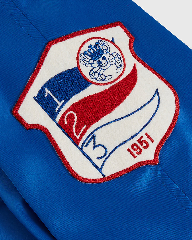 The Ebbets Field Santurce Cangrejeros Cotton Lined Satin Varsity Jacket features blue fabric with a red, white, and blue embroidered patch—a stylized bear, ball, 